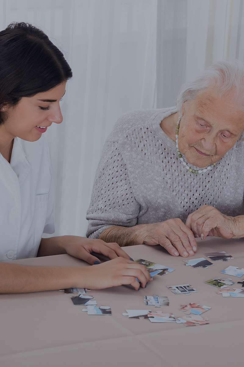 Young caregiver assisting an elderly woman in assembling a jigsaw puzzle at a table, promoting cognitive engagement and companionship.